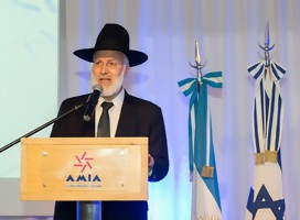 El rabino Davidovich haciendo uso de la palabra en un podio de oradores de AMIA. Vestido de negro, con sombrero negro. De fondo, la bandera de Israel y la de Argenitina.