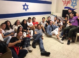 Un grupo de jóvenes sentados en el piso, y detrás, una bandera de Israel