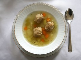 Un plato de sopa, sobre un plato llano. En la sopa, bolas de masa y verduras cortadas. A un costado, una cuchara